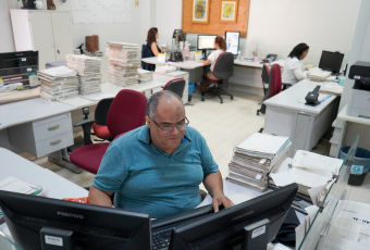 Há uma sala com pessoas sentadas trabalhando em frente aos seus computadores.