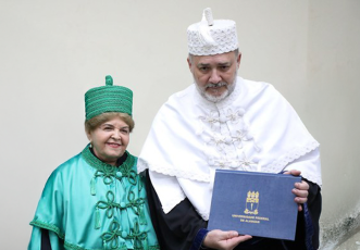 A imagem mostra duas pessoas vestindo trajes acadêmicos formais em uma cerimônia da Universidade Federal de Alagoas (UFAL). A mulher à esquerda veste uma beca verde com detalhes ornamentados e um chapéu correspondente, enquanto o homem  à direita usa uma beca branca e segura um diploma com o brasão da UFAL. O ambiente sugere um evento oficial de outorga de título ou formatura.