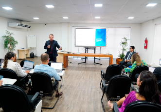 A imagem mostra uma sala de aula moderna onde está sendo ministrado um curso ou palestra. Um homem de terno, em pé, fala ao público utilizando um microfone, enquanto uma apresentação é exibida em uma tela grande ao fundo. Os participantes estão sentados em cadeiras com notebooks abertos, acompanhando o conteúdo. O ambiente é climatizado, bem iluminado e conta com decoração simples, com plantas e móveis de madeira.