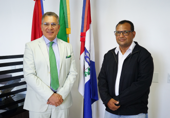 Há duas pessoas em pé, lado a lado, em um ambiente interno. Ao fundo, há três bandeiras. À esquerda, está um homem de cabelos curtos grisalhos e óculos. Ele veste um terno verde-claro com gravata e lenço de bolso em um tom de verde mais vibrante, além de camisa clara. Está sorrindo e com as mãos cruzadas à frente do corpo. À direita, está um homem, também usando óculos. Ele veste camisa branca, jaqueta preta e calça jeans clara. Está com expressão neutra e também com as mãos cruzadas à frente do corpo.