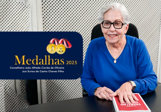 À direita da imagem, uma mulher, de cabelos curtos e brancos, veste uma blusa azul e usa óculos de armação escura. Ela está sorrindo e sentada à mesa, com as mãos apoiadas sobre um livro de capa vermelha. Usa um colar dourado e um relógio no pulso esquerdo. À esquerda da imagem, há um cartaz com fundo azul escuro.