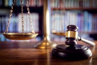 A imagem mostra uma composição simbólica do sistema judiciário: em primeiro plano, há um martelo de juiz (malhete) sobre uma superfície de madeira, indicando a autoridade das decisões judiciais. Ao fundo, levemente desfocada, aparece uma balança dourada da justiça, representando o equilíbrio e a imparcialidade do Direito. Estantes com livros jurídicos completam o cenário, reforçando o contexto de um ambiente jurídico ou tribunal.
