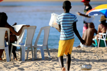 Menino negro de costas na areia de uma praia voltado para o mar, usando bermuda amarela e uma blusa azul de listras azuis escuros e segurando um depósito de comida na mão esquerda e, na direita, uma churrasqueira