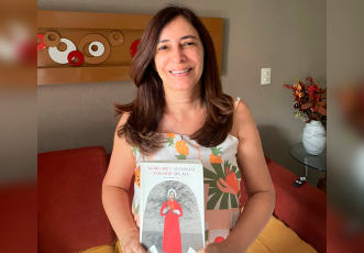 Mulher de cabelos castanhos lisos e longos sorri enquanto segura o livro O Conto da Aia, de Margaret Atwood. Ela está sentada em um sofá, com um vaso de flores e uma mesinha ao fundo.