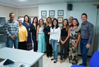 A imagem mostra um grupo de pessoas em pé, lado a lado, sorrindo para a câmera dentro de uma sala de trabalho. Há placas de premiação na parede ao fundo e mesas no primeiro plano. O ambiente é claro e organizado.