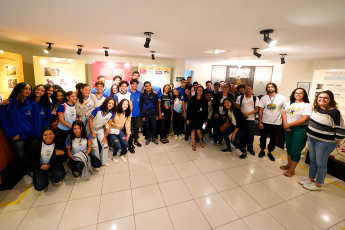 Grupo de estudantes e adultos posa sorrindo.Há no local, painéis informativos ao fundo. Estão em um ambiente interno iluminado, com piso branco e clima de visita educativa.