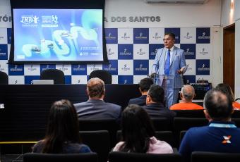 Homem alto de terno azul claro à frente do público do lado direito; à direita dele, um telão com o fundo azul claro
