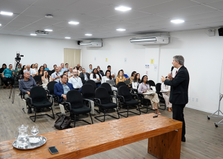 Homem alto de terno preto com o microfone na mão direita e em pé ministrando curso para uma plateia sentada.