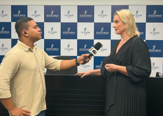 Entrevista em ambiente interno: repórter da TV Gazeta, homem pardo de cabelo curto e escuro, entrevista a juíza Estefânia Fernandes, mulher branca, loira, de cabelos lisos e médios, diante de painel do TRT-AL. Ambos estão de frente um para o outro em posição formal.