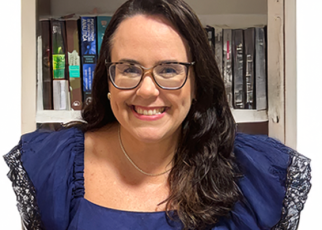 Mulher de pele clara, cabelos castanhos escuros e longos, está sorrindo. Ela usa óculos de grau e um vestido azul escuro com detalhes rendados nas mangas. Ao fundo, há uma estante com livros.