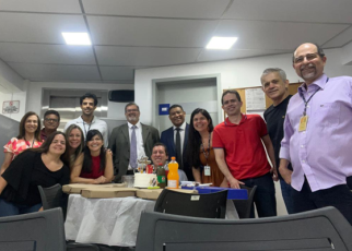 Grupo de pessoas sorrindo em uma confraternização, em volta de uma mesa com pizza, refrigerantes e bolo, em ambiente de trabalho.