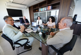 A foto mostra cinco pessoas reunidas em torno de uma mesa de vidro em um ambiente corporativo. Duas delas, em primeiro plano, vestem ternos claros e parecem estar em diálogo. Os outros três participantes, ao fundo, estão atentos e sorrindo, com notebooks e anotações sobre a mesa. O clima é de reunião formal, porém descontraída.