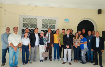 A imagem mostra um grupo de pessoas posando para uma foto em um ambiente interno, diante de uma parede clara com janelas e uma porta em arco. O grupo é diverso, composto por homens e mulheres, alguns de terno, outros com roupas casuais e crachás. Eles estão sorrindo.
