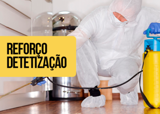 Pessoa vestida com macacão branco de proteção, luvas azuis e máscara realiza serviço de dedetização em um ambiente interno, ajoelhada no chão. Ela segura um equipamento de pulverização de cor amarela com detalhes em azul, utilizado para aplicar o produto. À esquerda da imagem, há um retângulo amarelo com o texto em preto: "Reforço Dedetização".