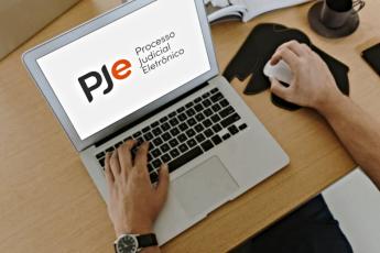 A imagem apresenta um computador com duas mãos digitando na tela a palavra PJe