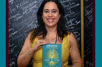 Há uma mulher  sorridente de cabelo escuro . Ele veste uma blusa amarela e segura um alivro de capa  esverdeada.