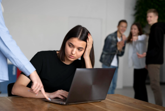 A imagem mostra uma mulher sentada à mesa, usando um notebook, com expressão triste e preocupada. Ela apoia uma das mãos na cabeça enquanto uma pessoa ao lado aponta algo na tela do computador. Ao fundo, três pessoas estão em pé, rindo e apontando em sua direção, sugerindo uma situação de constrangimento ou assédio no ambiente de trabalho.