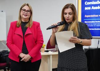 Há duas mulheres de peles claras. A primeira veste um terno vermelho e calça preta. Já a segunda, veste um vestido escuro e está segurando um microfone e um papel. 