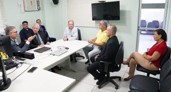 Grupo de pessoas participa de uma reunião em uma sala administrativa do TRT-AL, com integrantes sentados ao redor de uma mesa enquanto um homem de terno conduz a conversa. O ambiente conta com computadores, televisão e cadeiras distribuídas pelo espaço.