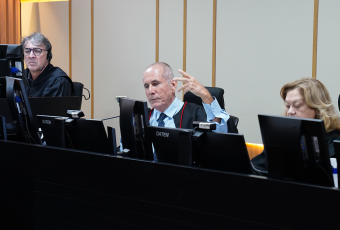 A imagem mostra três magistrados durante uma sessão de julgamento no plenário do Tribunal. Ao centro, um desembargador faz uso da palavra enquanto gesticula com a mão, demonstrando participação ativa nos debates. Os demais integrantes acompanham os trabalhos em seus respectivos postos, diante de monitores de computador, em ambiente formal e institucional.