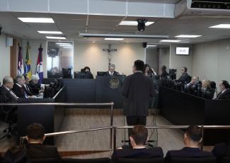 Fotografia colorida do plenário de um tribunal durante uma sessão de julgamento. Em primeiro plano, aparecem parcialmente as costas de algumas pessoas sentadas na plateia. Ao centro da imagem, um homem de terno escuro está de pé, de costas para a câmera, diante de um púlpito transparente, aparentemente realizando uma sustentação oral.  Ao fundo, os magistrados estão distribuídos em bancadas escuras dispostas em formato de "U". No centro da composição, atrás de uma bancada elevada com o brasão da República, 