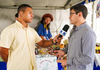 Há dois homens, um de camisa amarela entrevistando com microfone e outro de camisa xadrez sendo entrevistado. O ambiente é uma feira junina. Há uma mulher feirante atrás dos homens.