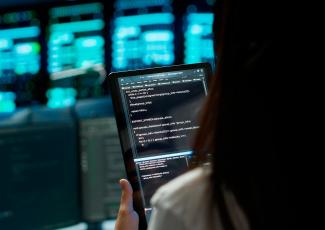 Uma pessoa é vista de costas segurando um tablet que exibe uma tela com código de programação. Ao fundo, há monitores desfocados com interfaces digitais em tons de azul e verde, compondo um ambiente tecnológico que remete a um centro de processamento de dados ou sala de monitoramento de sistemas.