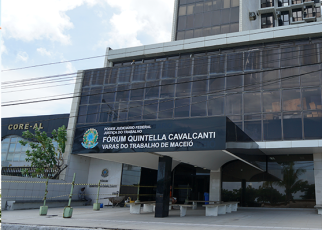 A foto exibe a fachada do prédio do Fórum Quintella Cavalcanti, onde funcionam as Varas do Trabalho de Maceió. O edifício possui estrutura moderna com amplas janelas espelhadas de tom escuro e a identificação oficial na entrada. Na parte inferior, vê-se a recepção coberta, bancos de espera na calçada e uma área demarcada por fitas zebradas.