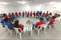 Várias pessoas vestidas com calça jeans, camisas nas cores zul, vermelha e branca, sentadas em cadeiras brancas dispostas em um círculo.