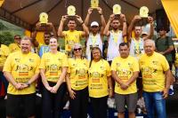 Grupo de pessoas vestidas de camisetas amarelas. O grupo da parte que tá mais elevada erguer troféus.