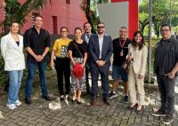 A imagem mostra um grupo de nove pessoas posando para uma fotografia em uma área externa arborizada. Os participantes estão alinhados lado a lado, usando trajes que variam entre o casual e o social, diante de um edifício de fachada avermelhada. Ao fundo, há jardins, árvores e uma estrutura vertical vermelha, compondo um ambiente institucional e acolhedor. A cena transmite um clima de integração e registro de um encontro ou atividade profissional.