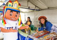 Há um mascote em primeiro plano da imagem. Ele é do TRT-AL. Em frente a estaõ duas mulheres em suas bancas de feira, onde estão exposta suas mercadoria. Uma deles está de chapéu de couro, e sorrir.