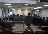 Fotografia colorida do plenário de um tribunal durante uma sessão de julgamento. Em primeiro plano, aparecem parcialmente as costas de algumas pessoas sentadas na plateia. Ao centro da imagem, um homem de terno escuro está de pé, de costas para a câmera, diante de um púlpito transparente, aparentemente realizando uma sustentação oral.  Ao fundo, os magistrados estão distribuídos em bancadas escuras dispostas em formato de "U". No centro da composição, atrás de uma bancada elevada com o brasão da República, 