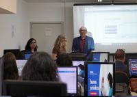 HÁ DUAS MULHERES E UM HOMEM DE PÉ FALANDO PARA VÁRIAS PESSOAS SENTADAS EM UMA SALA DE AULA, COM COMPUTADORES À SUA FRENTE.