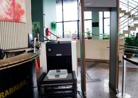 A foto mostra a entrada de um edifício com um posto de recepção e um sistema de controle de acesso. Em destaque, há um portal detector de metais e um equipamento de inspeção por raios X para bagagens, indicando um ambiente com medidas de segurança. Ao fundo, aparecem escadas, grandes janelas, plantas ornamentais e algumas pessoas circulando pelo local.