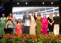 Na imagem, um grupo de autoridades, convidados e participantes posa para uma fotografia sobre o palco do Baile Estadual da Inclusão. Ao centro, um homem de terno cinza e gravata vermelha segura o certificado do Selo da Inclusão, ladeado por mulheres em trajes de gala e por dois participantes em cadeiras de rodas. Ao fundo, há um grande painel de LED exibindo uma imagem do evento, enquanto refletores iluminam o palco. Na parte frontal, um arranjo com flores tropicais vermelhas e folhagens ornamenta o cenário