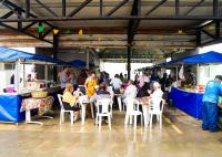 Há um área grande cobereta e várias barracas de feira. Também há mesas e cadeiras dispostas no local com pessoas sentadas.