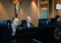 Uma sessão de julgamento no plenário do TRT-AL, com três magistrados sentados à bancada. Ao centro, o desembargador Antônio Catão sorri para a câmera, enquanto os demais participantes acompanham os trabalhos. Ao fundo, destaca-se um crucifixo fixado na parede revestida em madeira e um monitor indicando o horário da sessão.
