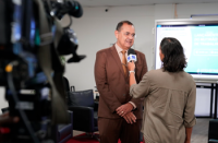 Uma pessoa concede entrevista a uma repórter em um ambiente interno. A jornalista segura um microfone em direção ao entrevistado, enquanto uma câmera de televisão aparece em primeiro plano, registrando a gravação. Ao fundo, há um monitor exibindo a identidade visual de um evento, reforçando o contexto institucional da entrevista.