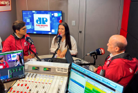 "Entrevista em estúdio de rádio, com três participantes sentados diante de microfones durante a transmissão de um programa da Pan News. Ao centro, uma mulher fala enquanto é entrevistada por dois apresentadores, em um ambiente equipado com mesa de áudio, monitores e computadores."