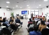 Auditório com várias pessoas sentadas. Elas assitem uma pessoa falar. Há um telão atrás da mulher que fala.