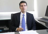 Há um homem de terno preto e gravata lilás, sentado e trabalhando em uma mesa com computador