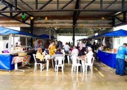 Há um área grande cobereta e várias barracas de feira. Também há mesas e cadeiras dispostas no local com pessoas sentadas.