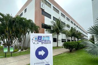 Em primeiro plano, o banner nas cores azul e branco, com um círculo no meio e nele inserido uma seta de indicação na cor branca e, fora a círculo, a frase mutirão vaga inclusiva de trabalho; ao fundo, a fachada da Faculdade Unit.