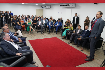 À direita e em pé, homem alto, moreno, de cabelos pretos,  usando óculos, terno cinza escuro, gravata vermelha e com o microfone na mão direita, à esquerda e ao fundo, diversas pessoas, homens e mulheres, assistindo à apresentação. Os homens vestindo ternos escuros e as mulheres com vestidos e roupas sociais.
