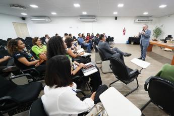 Homem de terno cinza, cabelos grisalhos em pé, falando em frente a homens e mulheres sentados em um auditório