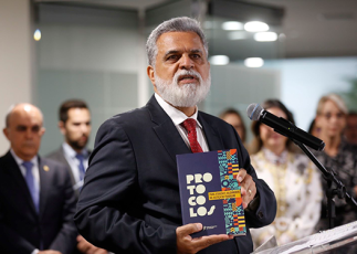 Homem de barba e cabelos brancos vestindo terno cinza e gravata vermelha segura um livro enquanto fala ao microfone