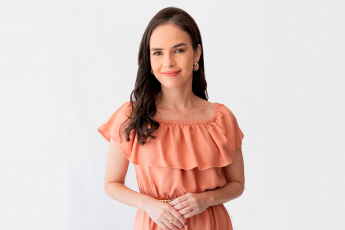 Mulher jovem de pele clara e cabelos longos escuros. Ela veste um vestido de cor clara e tá  posicionada para uma foto.