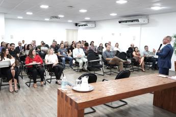 Há um homem de pé segurando um microfone falando para uma plateia sentada.. Ele usa óculos e veste terno escuro. 