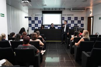 Homem de terno preto e camisa branca em pé do lado direito do auditório falando para várias pessoas que estão sentadas de frente para ele.